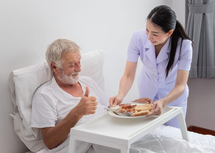 Happy nurse serving elderly senior man breakfast on bed in bedroom at nursing home, thumb up
